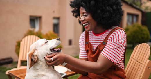A person wearing overalls and a striped shirt pets a light-colored dog in a backyard setting.
