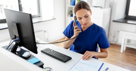 A person in blue scrubs is sitting at a desk with a computer and a phone,